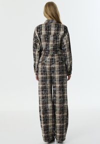 Braun-beige karierter Overall mit langen Ärmeln, dehnbarem Bund und weit geschnittenen Hosenbeinen, aus einem strukturierten Stoff gefertigt. Rückansicht gezeigt.