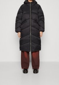 Svart puffjacka med huva, med ett chevron-quiltat mönster och dragkedja fram. Kombineras med vida röda denimbyxor och svarta skor.