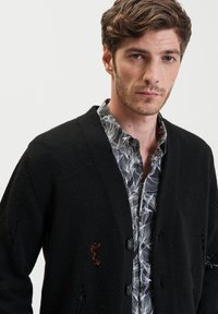 Zwarte gebreide cardigan met een V-hals, vier knopen, kleine uitsparingen en een borduursel, gelaagd over een bloempatronen shirt.