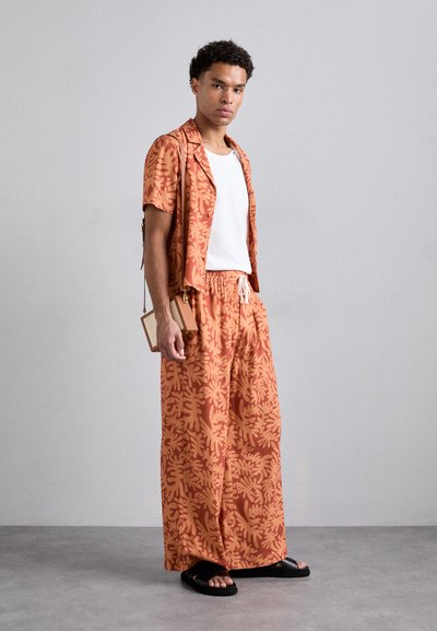 Short-sleeve patterned shirt and wide-leg pants in orange hues with leaf motifs. Paired with black slide sandals and a small crossbody bag.