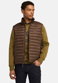 Bruine puffer vest met horizontaal gestikt patroon, hoge kraag en voormet rits, gedrapeerd over een groene sweatshirt met grafisch ontwerp.