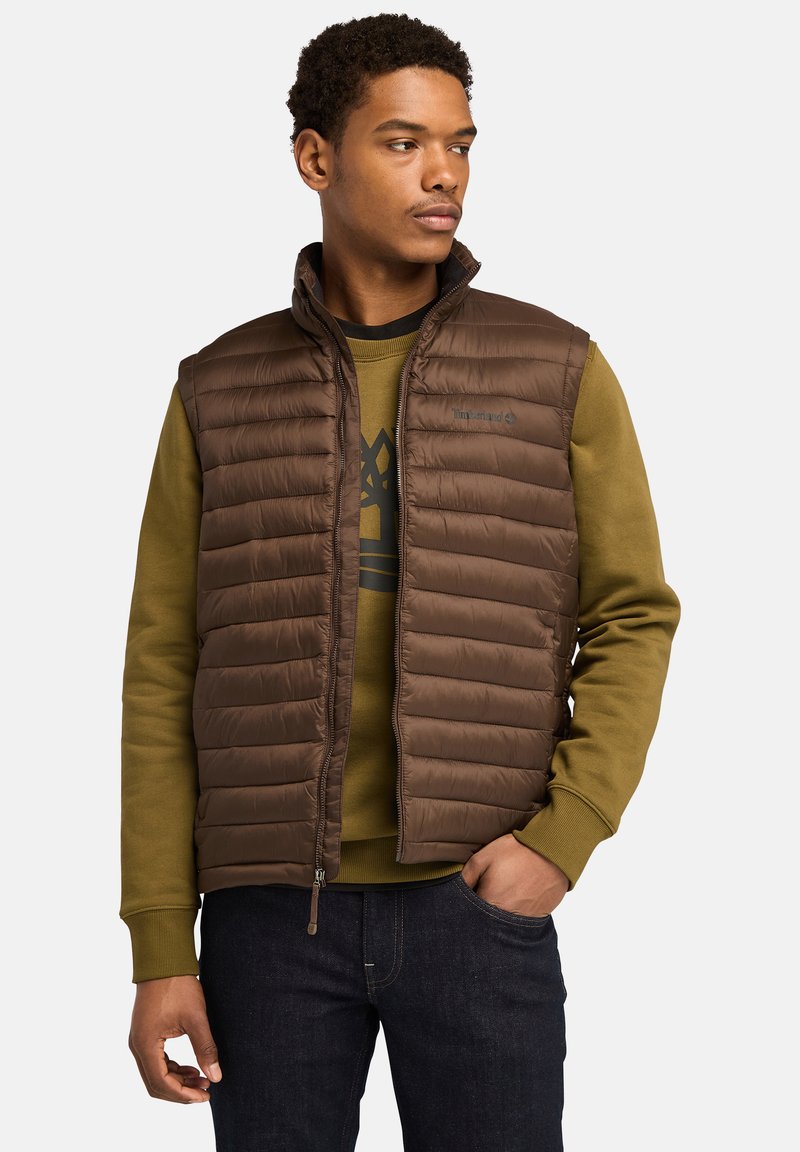 Bruine puffer vest met horizontaal gestikt patroon, hoge kraag en voormet rits, gedrapeerd over een groene sweatshirt met grafisch ontwerp.
