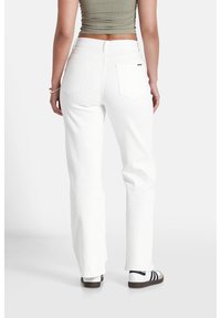 Persona de espaldas, vistiendo una blusa verde ajustada y jeans blancos. Se encuentran ante un fondo liso, mostrando la parte posterior del atuendo.