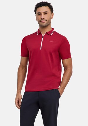 Mann trägt rotes Poloshirt mit kurzen Ärmeln, weißem Reißverschluss und Kragenstreifen, kombiniert mit dunkler Hose, steht mit einer Hand in der Tasche.