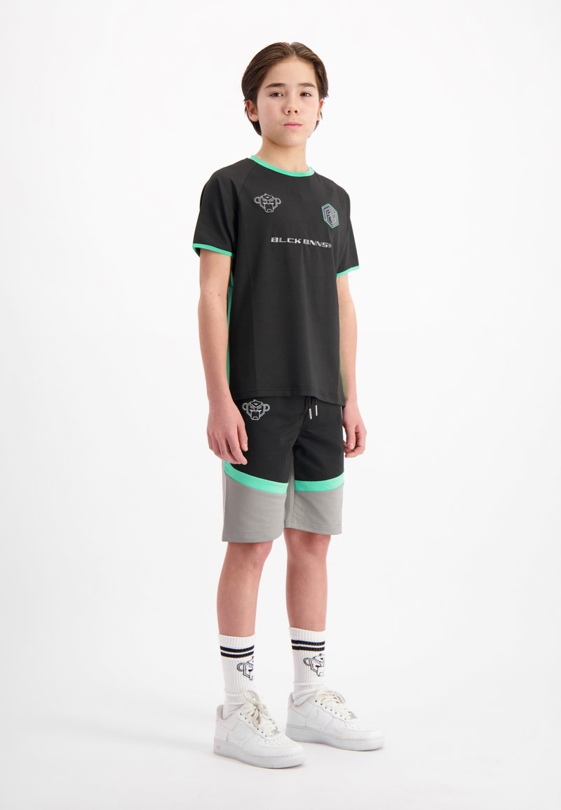 Zwart sport T-shirt met groene accenten, logo op de borst, gecombineerd met zwart-grijze shorts met groene bies, witte sneakers en gestreepte sokken.