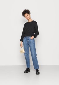 Black long-sleeve top with gathered waist, blue straight-leg jeans, black platform shoes, and a small yellow handbag. Neutral background.