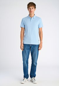 Polo blu chiaro con maniche corte e un piccolo logo, abbinato a jeans blu scuro e sneakers bianche. Design semplice e vestibilità casual.