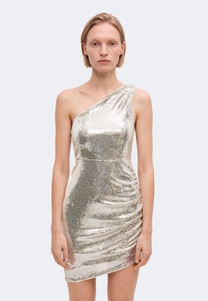 Une femme porte une mini-robe ajustée argentée à sequins, asymétrique et dégageant une épaule, sur un fond clair uni.