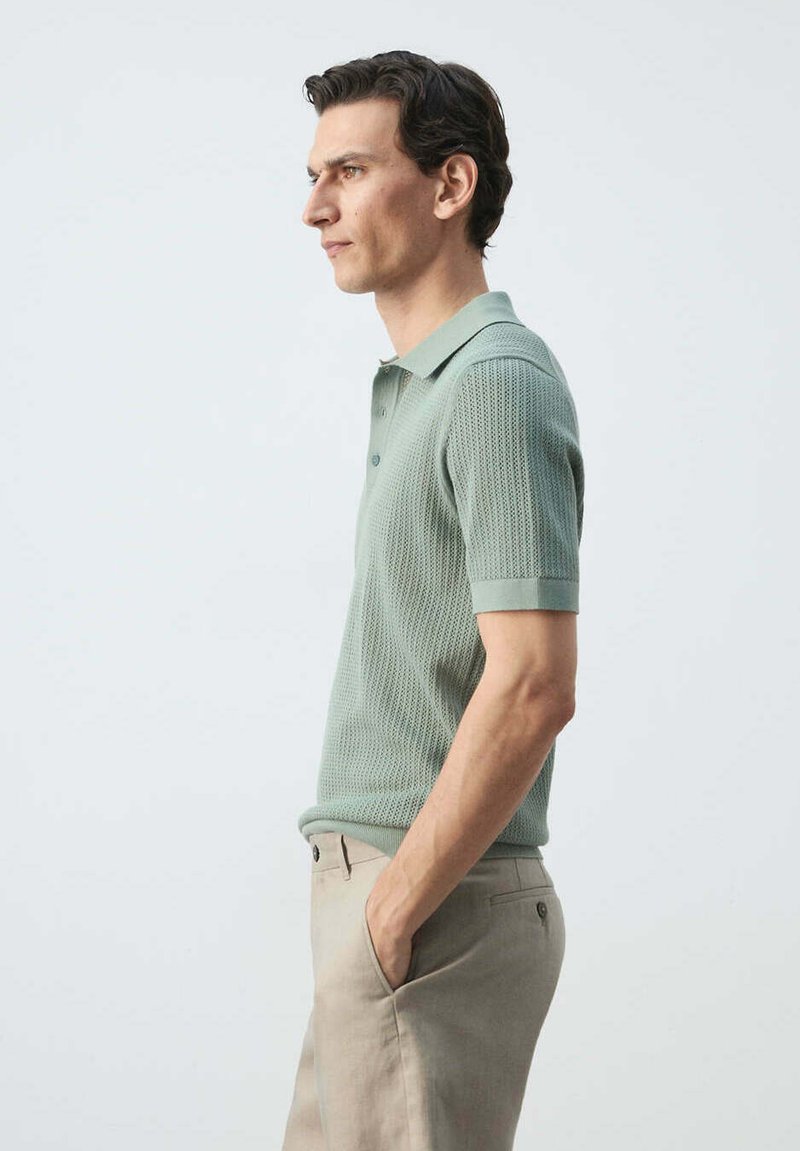 Homme en polo tricoté manches courtes vert clair et pantalon beige, debout de profil, mains dans les poches, sur fond uni.
