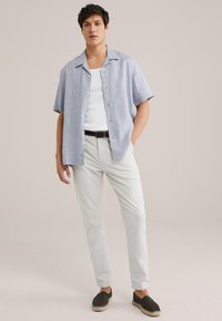 Camicetta azzurra chiara a maniche corte, canotta bianca, pantaloni grigi chiari, cintura intrecciata nera e scarpe scure senza lacci. Tessuto morbido, vestibilità casual.