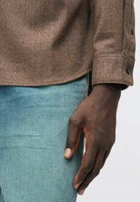 Καφέ υφασμάτινο πουκάμισο με κουμπωτές μανσέτες, φορεμένο πάνω από ανοιχτόχρωμα μπλε τζιν. Ορατό χέρι με φυσικό τόνο δέρματος που resting πάνω από τα τζιν.