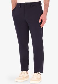 Pantaloni slim fit blu navy con tasche laterali, chiusura con bottone e orli risvoltati, abbinati a sneakers beige. Tessuto dalla texture liscia.