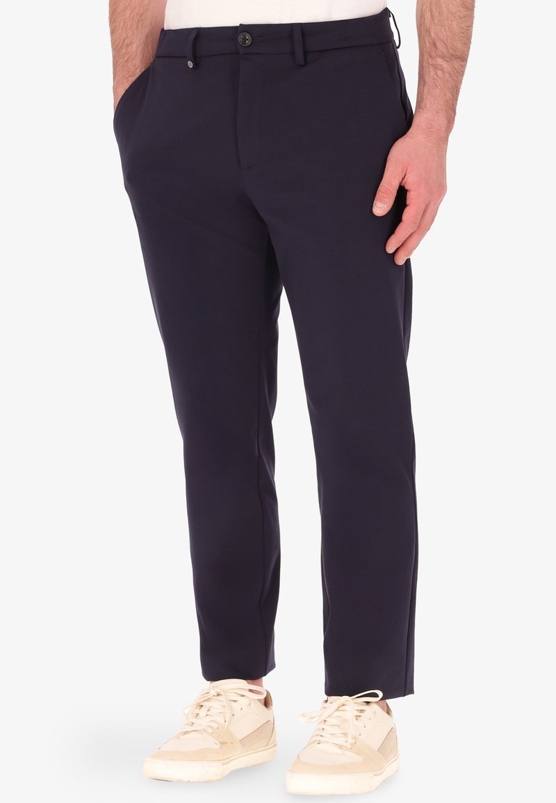 Pantaloni slim fit blu navy con tasche laterali, chiusura con bottone e orli risvoltati, abbinati a sneakers beige. Tessuto dalla texture liscia.