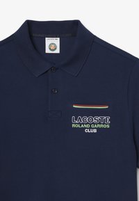 Marinblå piké med texturerad tyg, med en treknapppplacket och broderad "Lacoste Roland Garros Club"-logotyp.