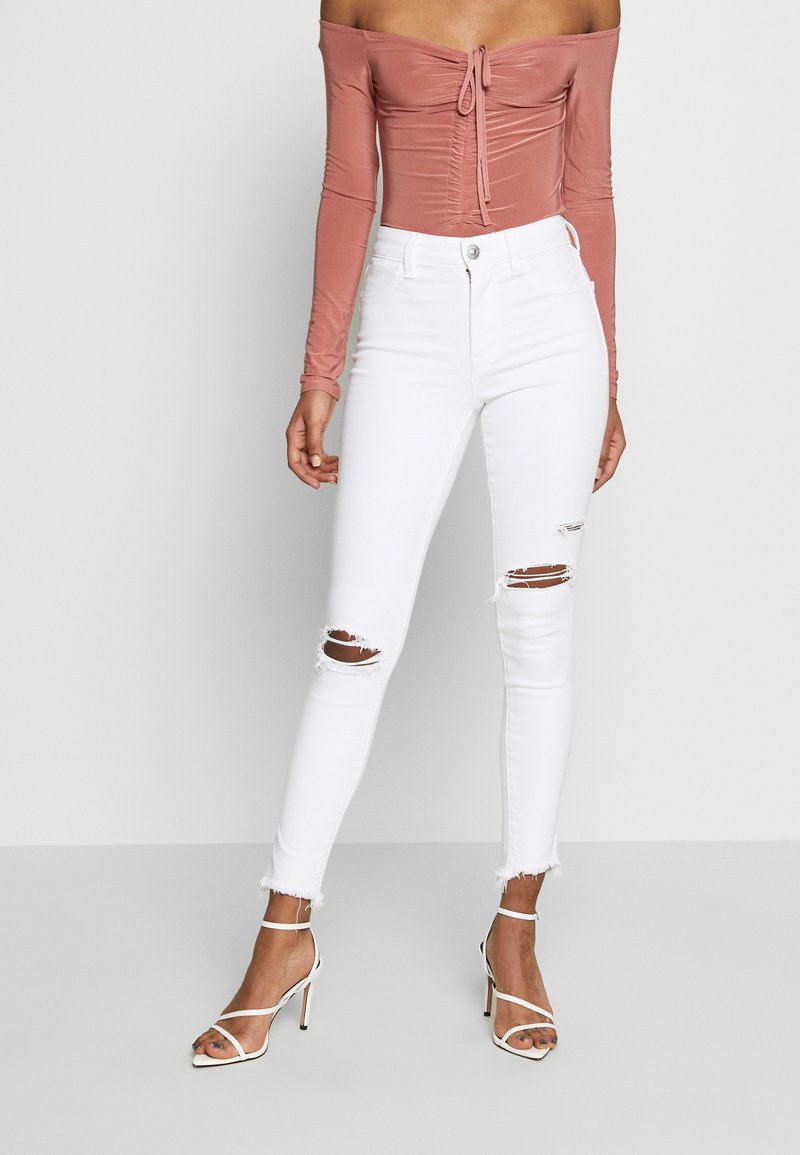 Haut rose clair à épaules dénudées avec un détail froncé, associé à un jean skinny blanc taille haute avec des genoux déchirés et des ourlets effilochés. Sandales à talons blanches.