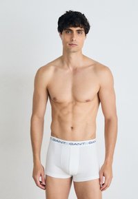 Weiße Boxershorts mit einem blauen elastischen Bund, der das "GANT"-Logo trägt. Glattes Material und schmaler Schnitt. Das Modell steht vor einem neutralen Hintergrund.
