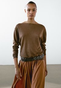 Hnědý pletený svetr s širokým lodním výstřihem, zkombinovaný s rustově zbarvenou plisé sukní. Oblečení je doplněno černým pásem s kulatými kovovými detaily.