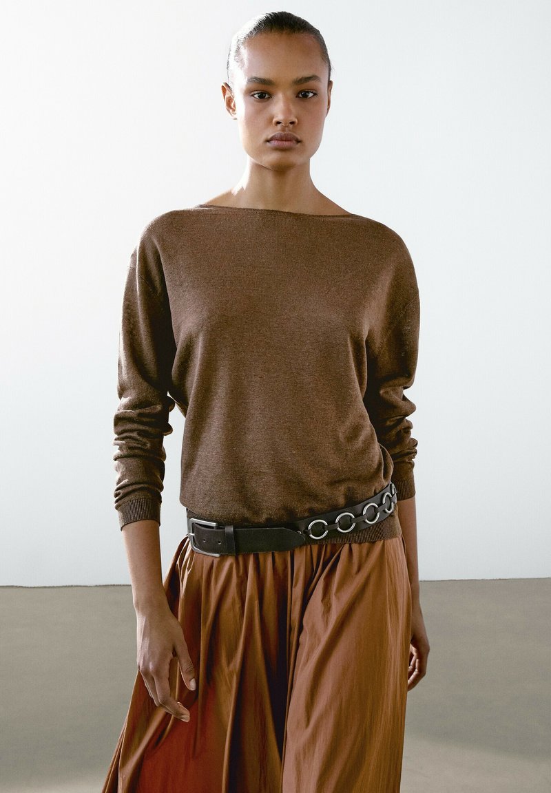 Hnědý pletený svetr s širokým lodním výstřihem, zkombinovaný s rustově zbarvenou plisé sukní. Oblečení je doplněno černým pásem s kulatými kovovými detaily.