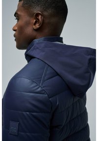 Granatowa ocieplana kurtka z usztywnionym kapturem, pikowanym wzorem i widocznymi detalami szwów. Posiada naszywkę z logo na rękawie.