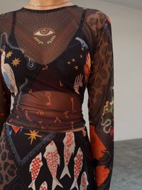 Μαύρο φόρεμα από διαφανές δίχτυ με ζωντανά, πολύπλοκα σχέδια ψαριών, χεριών και ουράνιων συμβόλων, με εφαρμοστή μέση και μακριά μανίκια.