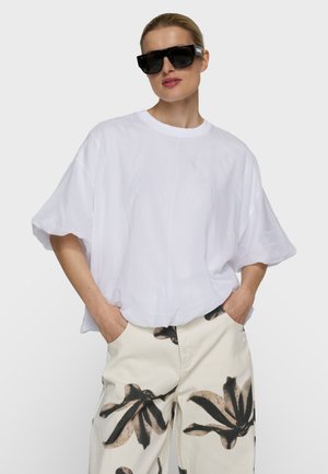 Frau trägt übergroßes weißes T-Shirt, helle Hose mit großem schwarzem Blumenmuster, schwarze rechteckige Sonnenbrille, Hände in den Taschen, neutraler Hintergrund.