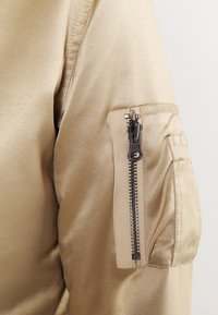 Giacca bomber in satin beige con tasca zip sul maniche, caratterizzata da una texture morbida e accessori metallici.