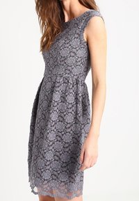 Robe en dentelle grise avec un corsage ajusté, des manches courtes et une jupe évasée. Motif floral remarquable et détails en bordure festonnée.