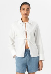 White long-sleeve shirt with a collar and front tie detail, featuring a relaxed fit and a partially open design, paired with denim shorts.