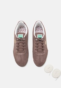 Sneakers de camurça castanha com solas brancas, apresentando um sistema de atacadores tradicional, colarinho acolchoado e palmilhas com padrão geométrico, acompanhados por atacadores brancos.