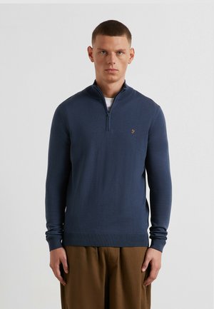 Pull-over en maille bleu marine avec col à fermeture éclair partielle, poignets et ourlet côtelés, arborant un petit logo sur la poitrine gauche, accompagné d'un pantalon marron.