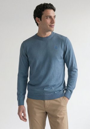 Mann mit dunklen lockigen Haaren, der einen blauen Langarmpullover und beige Hosen trägt, steht mit einer Hand in der Tasche und schaut zur Seite.