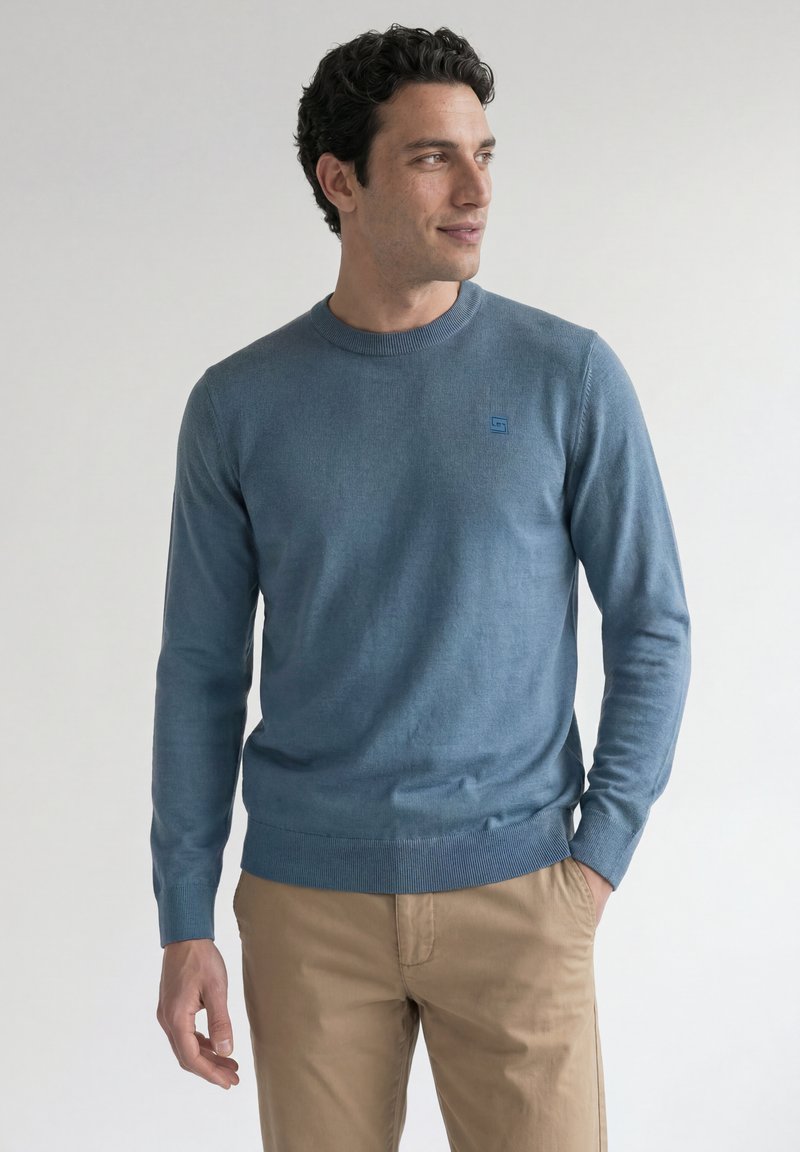 Homme aux cheveux bouclés et foncés portant un pull bleu à manches longues et un pantalon beige, debout, une main dans la poche, regardant sur le côté.