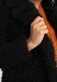 Sort, fuzzy jakke med blød tekstur, der har en løs pasform. En hånd griber fat i kraven og afslører en orange strikket sweater nedenunder.