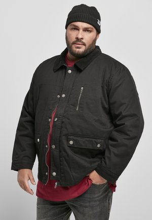 Jachetă neagră cu fermoar și închidere cu capse, buzunare laterale și pe piept, guler din catifea, purtată peste o cămașă roșie. Material texturat, croi lejer.