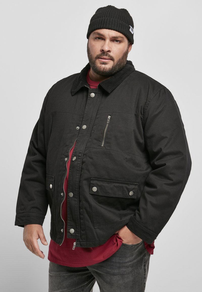 Giacca nera con chiusura a zip e bottoni a pressione, tasche laterali e sul petto, colletto in velluto a coste, indossata sopra una camicia rossa. Tessuto testurizzato, vestibilità comoda.
