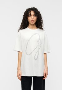 Λευκό βαμβακερό t-shirt με μεγάλο, λαμπερό ασημί γράμμα "G" σε καλλιγραφικό στιλ στο μπροστινό μέρος. Μανίκια κοντά και χαλαρή εφαρμογή. Μινιμαλιστικό στυλ.