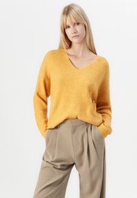 Maglione a costine giallo con scollatura a V profonda e maniche lunghe, abbinato a pantaloni beige a vita alta, mani in tasca.
