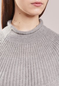 Pull gris clair en maille avec une texture côtelée et un col montant arrondi. Le tissu semble doux et chaud, offrant un aspect confortable.
