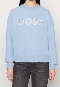 Ljusblå sweatshirt med ribbade ärmar och nederkant, med en texturerad vit "BOSS" grafik över bröstet. Mjuk material, avslappnad passform.