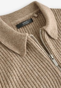 Brauner, gerippter Strickpullover mit einem Reißverschlusskragen und silbernem Reißverschluss. Das angeheftete Etikett trägt die Aufschrift "NEXT", was auf die Marke hinweist. Weicher, strukturierter Stoff.