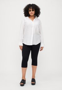 White long-sleeve shirt with a collar, button front, and tied cuffs paired with black capri leggings and black platform sandals.
