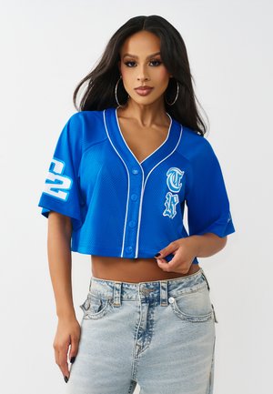 Frau mit langen dunklen Haaren trägt ein blaues, kurz geschnittenes Sporttrikot mit Knöpfen und weißen Buchstaben sowie hellen, hoch taillierten Jeans vor einem weißen Hintergrund.