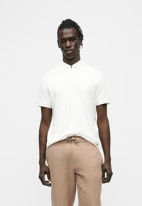 Homme aux dreadlocks courtes portant un polo blanc à fermeture éclair à manches courtes et un pantalon beige à cordon, debout devant un fond uni.