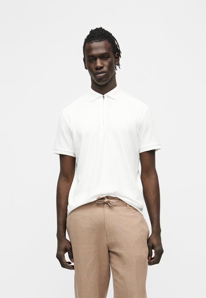 Homme aux dreadlocks courtes portant un polo blanc à fermeture éclair à manches courtes et un pantalon beige à cordon, debout devant un fond uni.