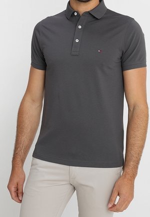 Mand iført en mørkegrå kortærmet polo med tre hvide knapper og lyse beige bukser, stående mod en ensfarvet baggrund.