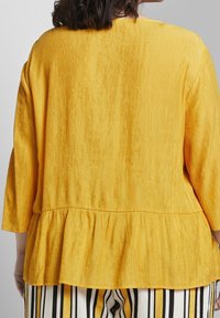 Top giallo a peplo in tessuto testurizzato, con maniche a tre quarti e orlo arricciato, abbinato a pantaloni a righe.