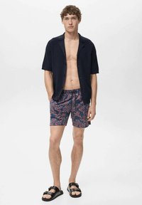 Majica s kratkimi rokavi v mornarsko modri barvi, kopalne hlače z vzorcem v koralni zasnovi v modri in rdeči barvi, v kombinaciji s črnimi sandali. Model stoji na enotni ozadju.