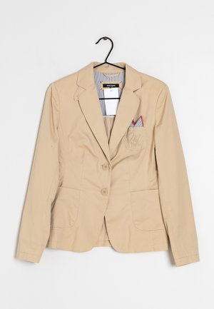 Beigefarbener maßgeschneiderter Blazer mit zwei Knöpfen, eingekerbtem Revers, zwei Vordertaschen, einer Brusttasche mit einem gefalteten Einstecktuch, hängend an einem schwarzen Kleiderbügel.