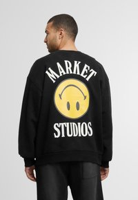 Fekete kapucnispulóver, nagyméretű sárga mosolygó arc grafikával, fehér szöveggel "MARKET" fent és "STUDIOS" lent.