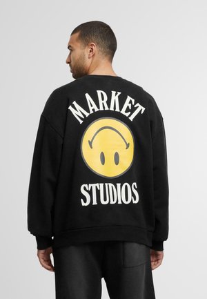 Sort sweatshirt med et stort gult smiley-ansigt grafisk design med teksten "MARKET" ovenfor og "STUDIOS" nedenfor i hvid.